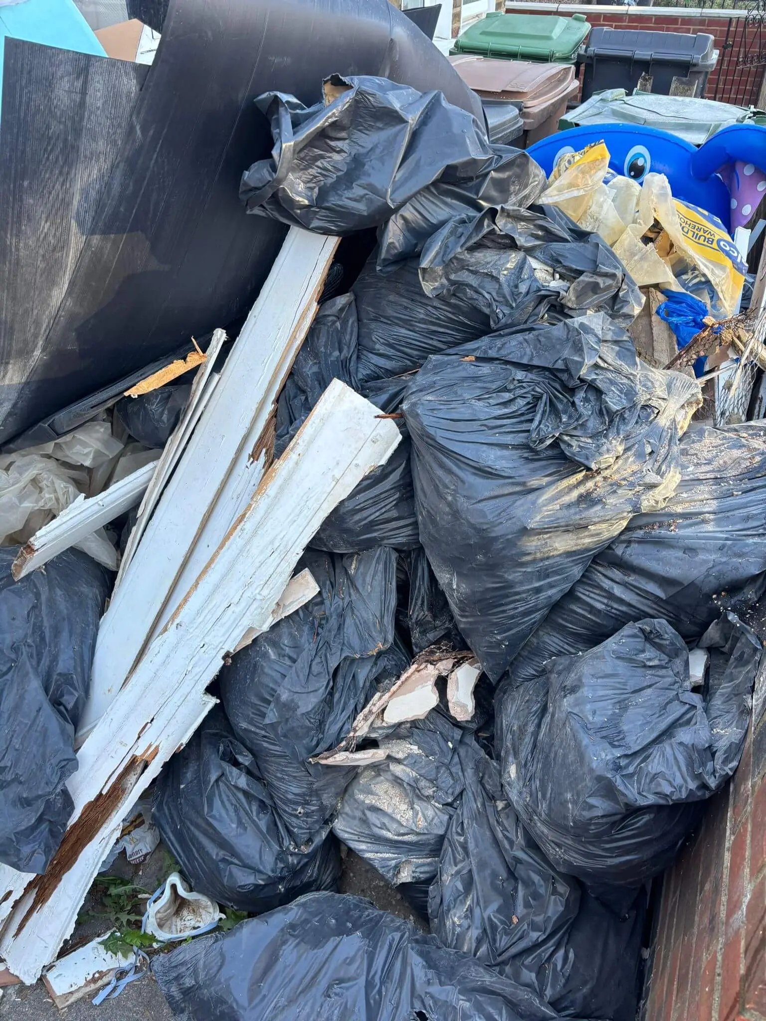 Bagged mixed waste, timber offcuts and lengths of skirting board stacked against a fence.