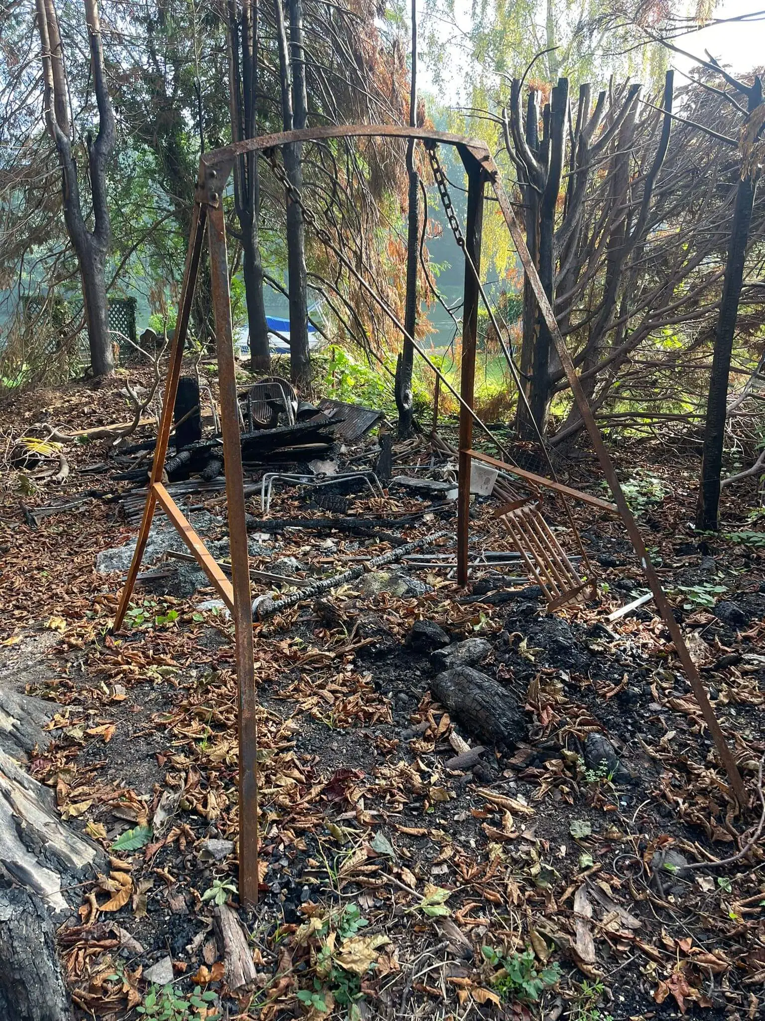 Fire-damaged garden with burnt trees, a rusted swing frame and charred furniture on scorched ground.