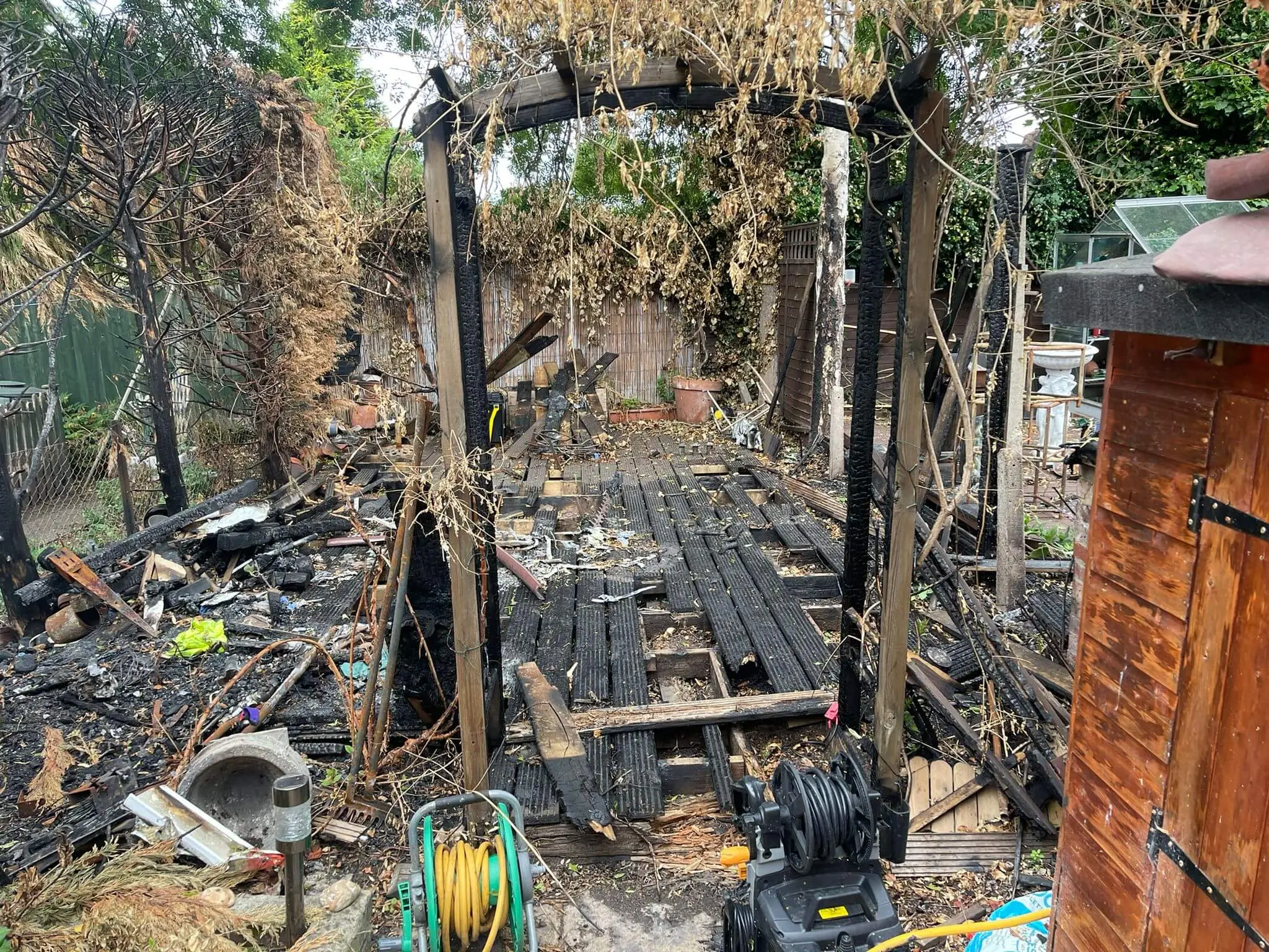 Charred decking and a burnt pergola with fire-damaged timber, hose reels and a pressure washer.