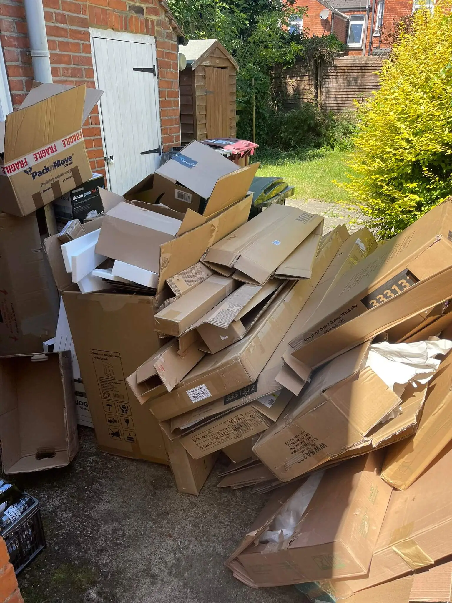 Flattened cardboard boxes, flat-pack packaging and polystyrene stacked against a garage door and shed.