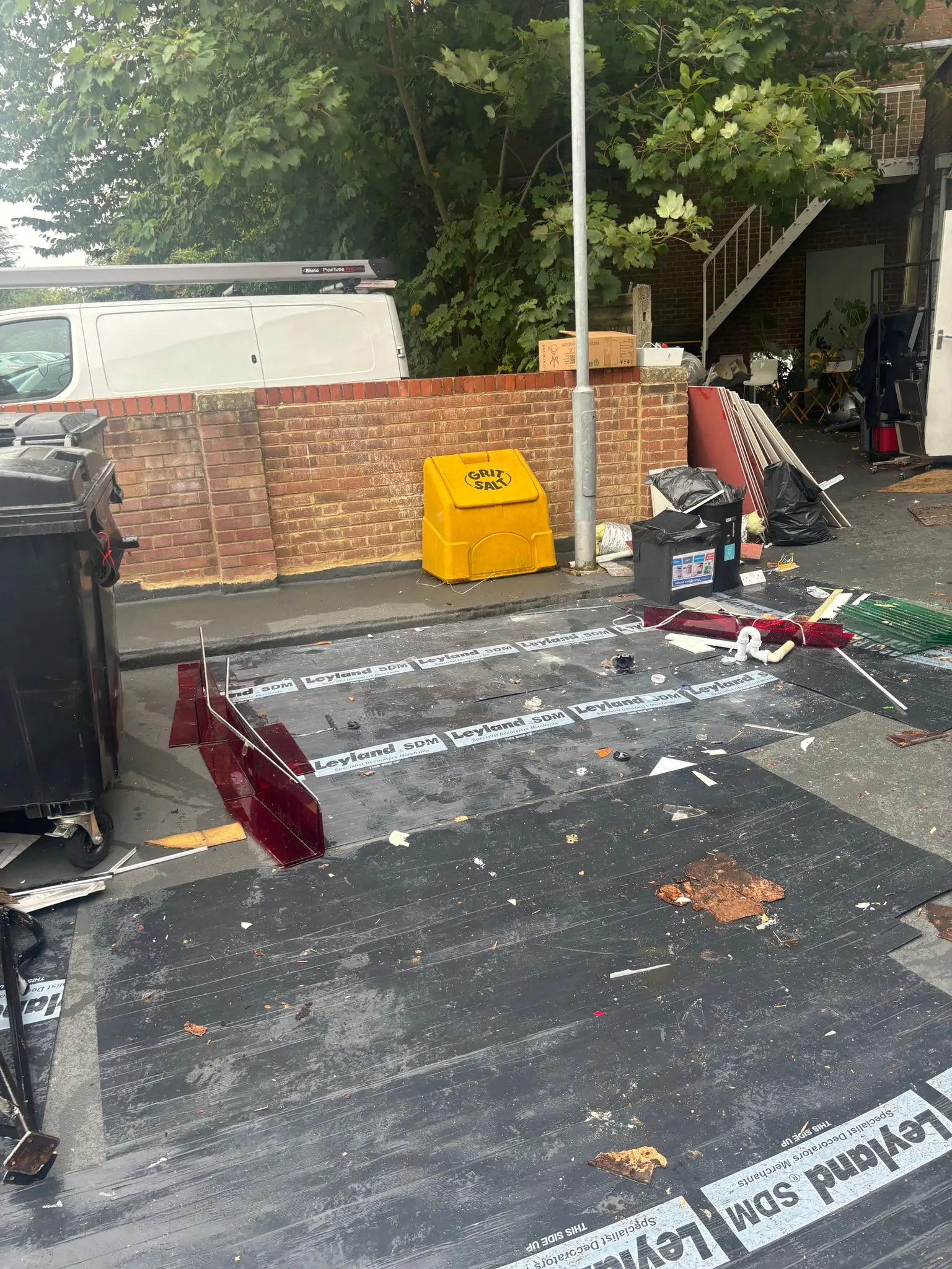 The same tarmac area with the pile removed, dust sheets laid down, a grit bin, wheelie bins and a few boards remaining.