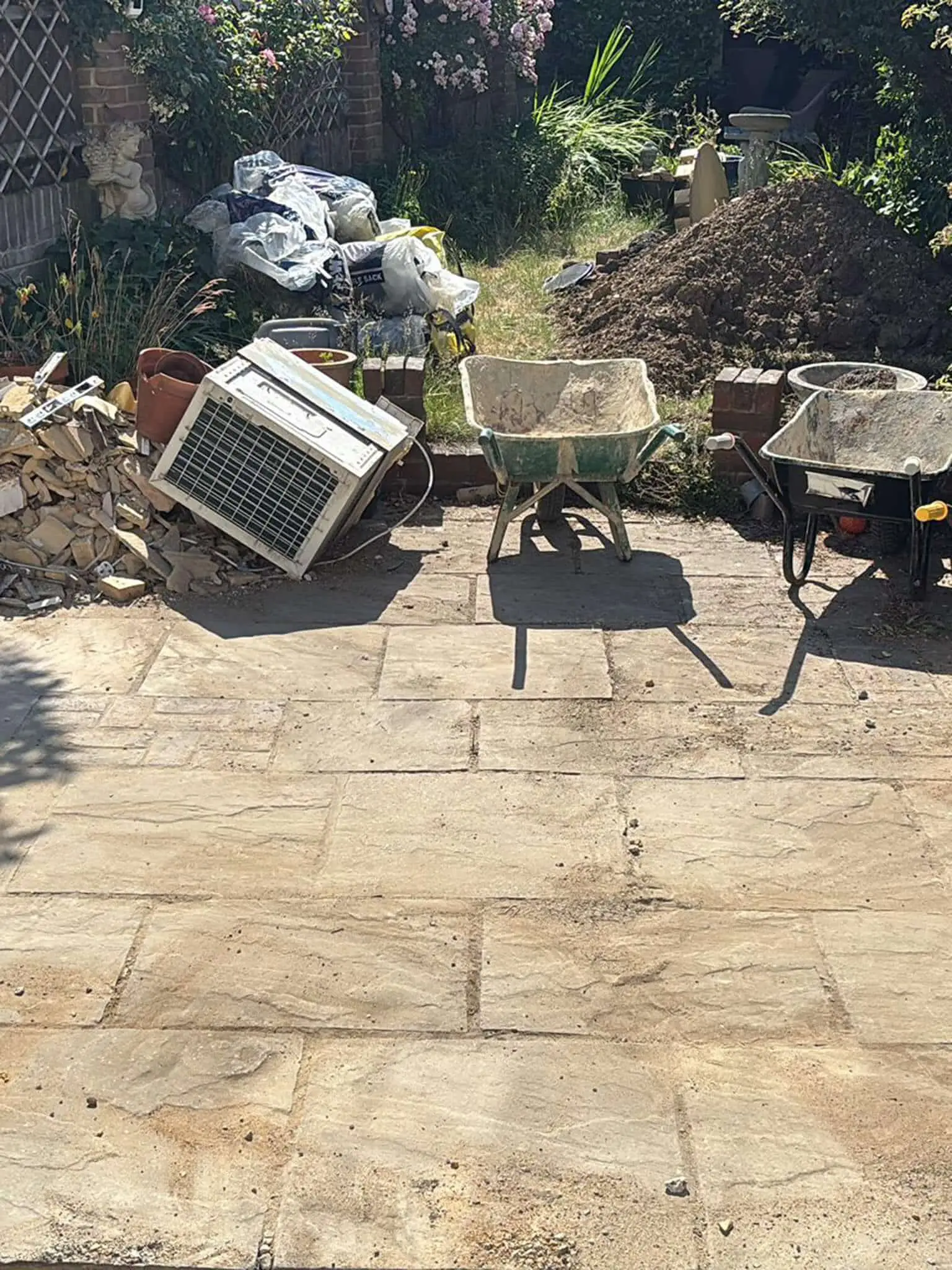 The same sandstone patio swept clear with wheelbarrows on it, a rubble pile, tonne bags and a soil heap still at the garden edge.