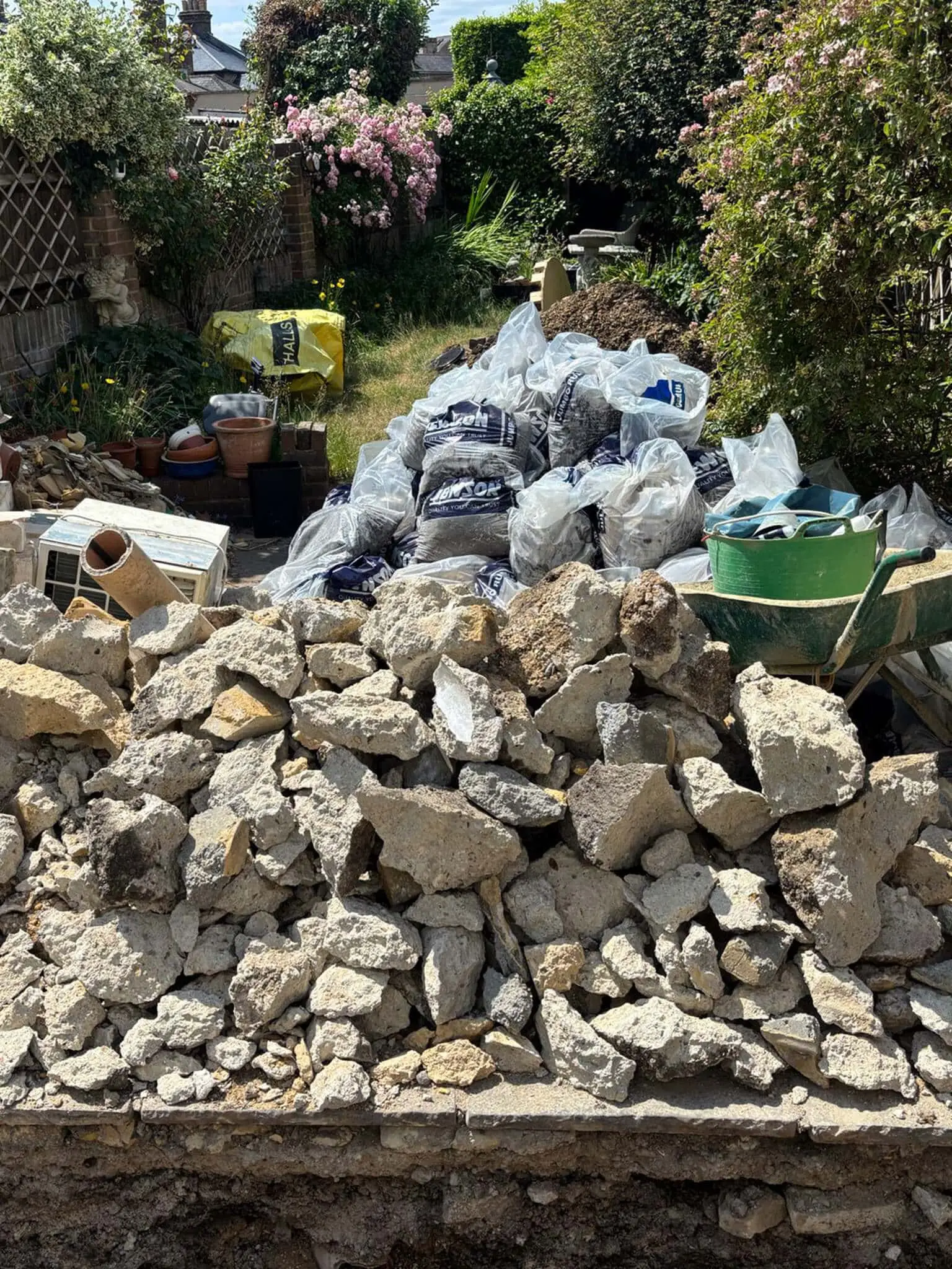 Broken concrete, bagged builders waste and a wheelbarrow gathered in a garden before clearance.