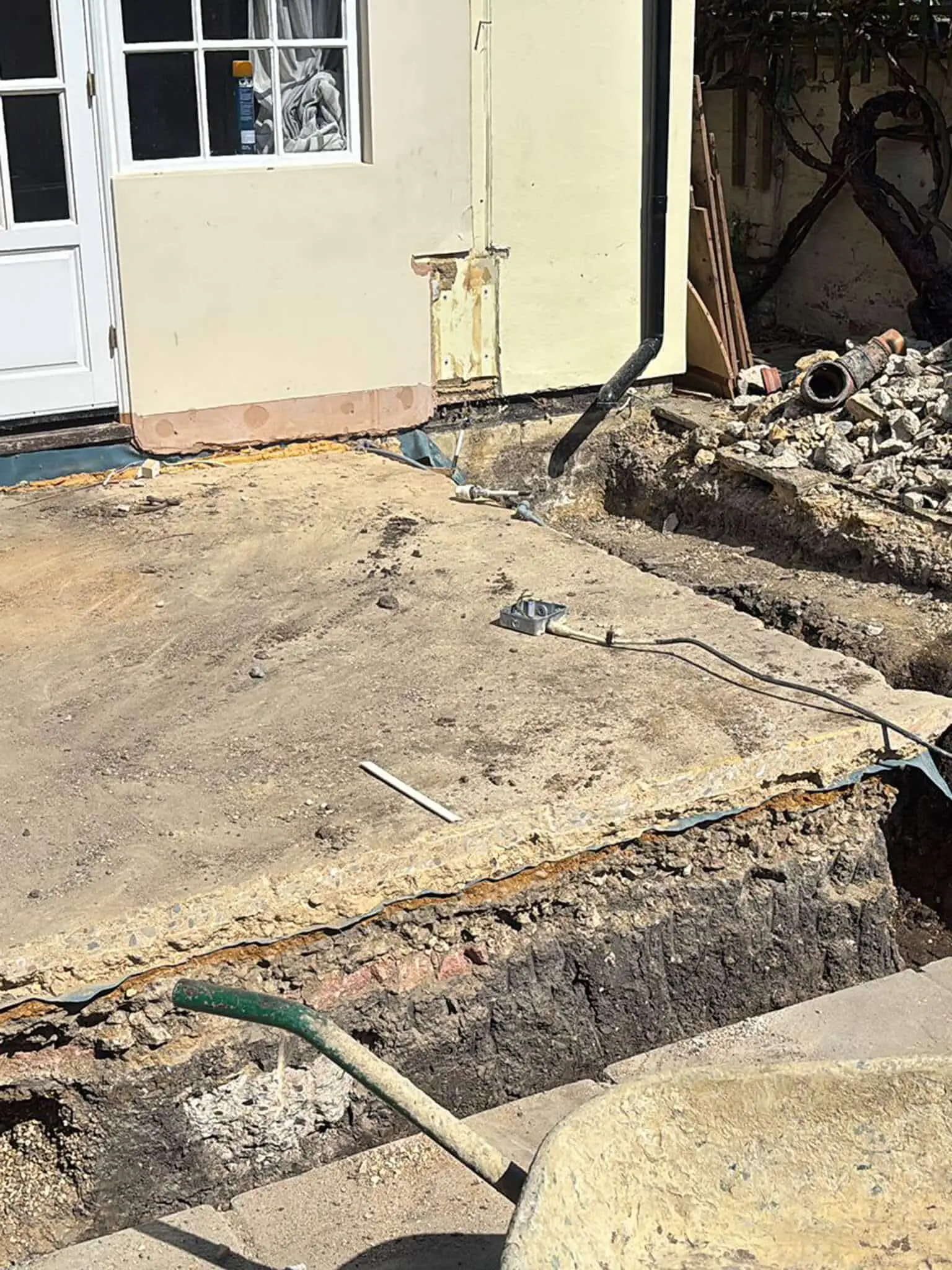 Excavated ground beside a house after the rubble sacks were taken, with an open trench, exposed pipework and a broken slab remaining.