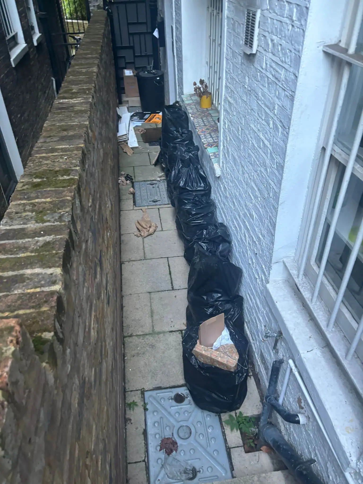 Narrow passage between two buildings filled with tied black sacks, cardboard and a bin at the far end.